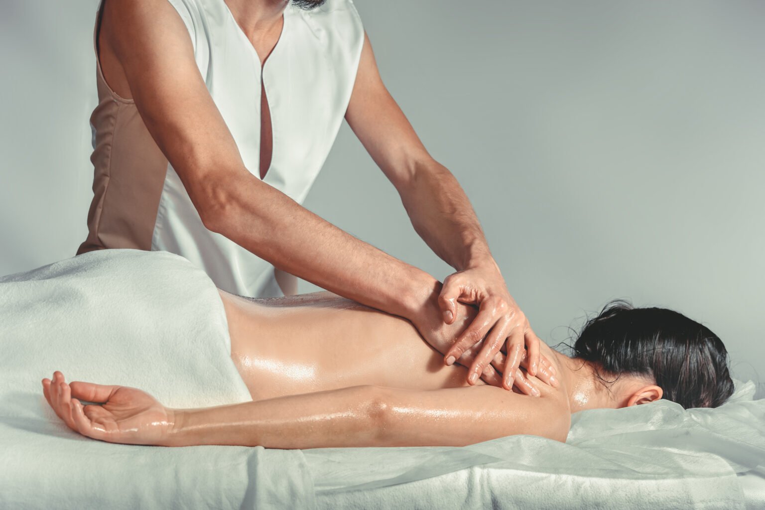 Massage oil. A young professional male masseur makes Thai massage. Patient woman is lying down on a bed and is covered with white towel. Treatment, rest, relaxation, spa, health care, medicine